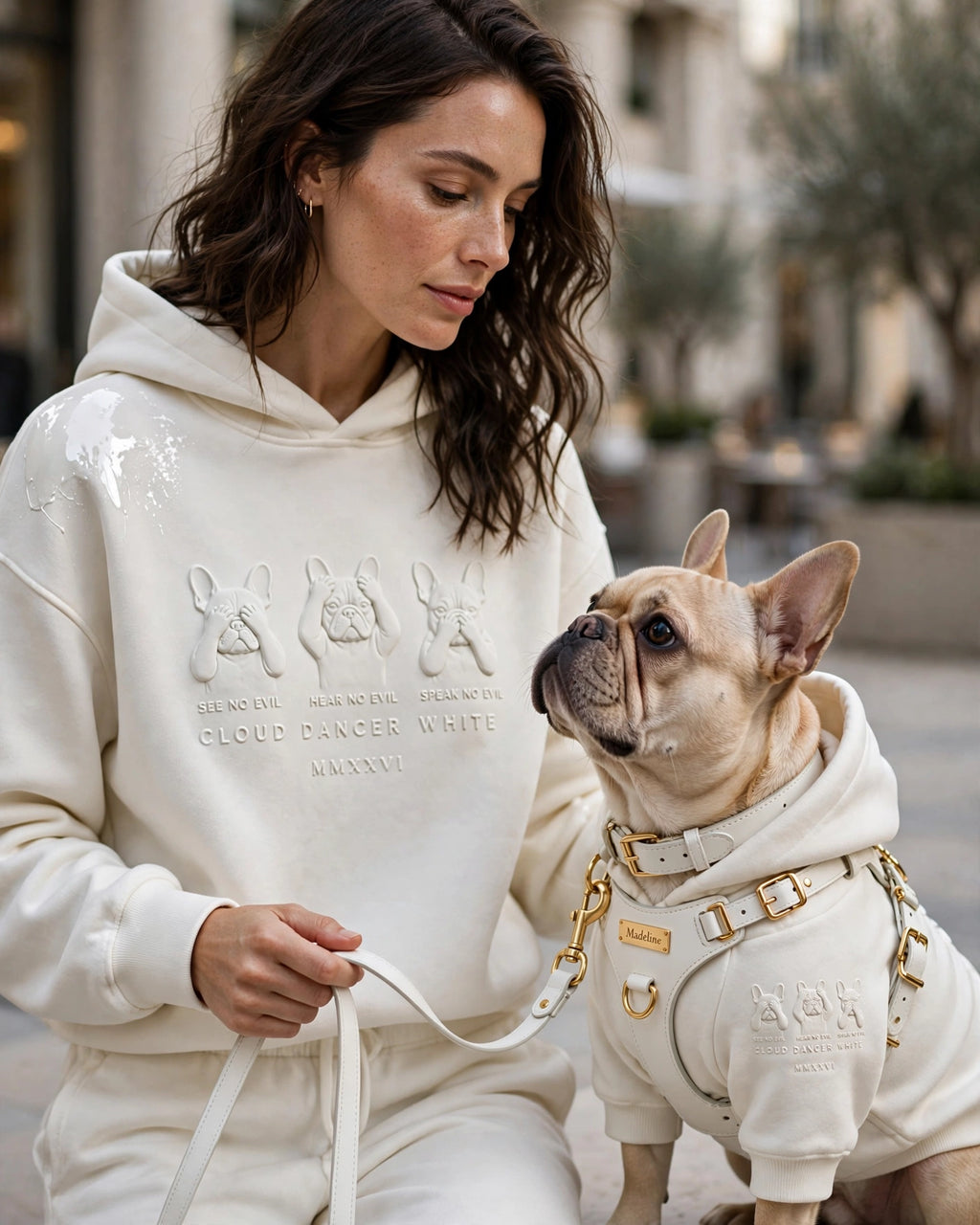 A woman and a French Bulldog wearing matching off-white hoodies and matching pants in an outdoor urban setting. The hoodies feature embossed rabbit graphics and text including 'MERCER ARROWS CLAUDIA WHITE CLOUDancer WHITE'.
