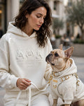 A woman and a French Bulldog wearing matching off-white hoodies and matching pants in an outdoor urban setting. The hoodies feature embossed rabbit graphics and text including 'MERCER ARROWS CLAUDIA WHITE CLOUDancer WHITE'.