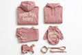 A coordinated pet apparel set in dusty pink, including a hoodie, sweatpants, a small hoodie, a harness, a collar, and a leash, all arranged on a white background. The hoodies and small hoodie feature the words 'Doggy Style Society' in a cream script.