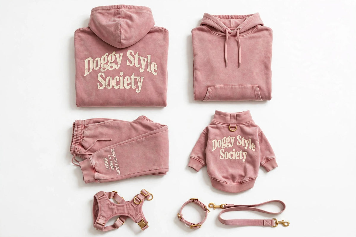 A coordinated pet apparel set in dusty pink, including a hoodie, sweatpants, a small hoodie, a harness, a collar, and a leash, all arranged on a white background. The hoodies and small hoodie feature the words 'Doggy Style Society' in a cream script.