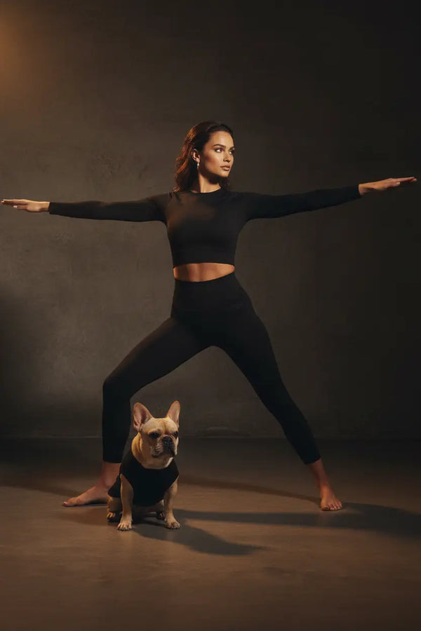 A woman in a black activewear set poses in a yoga warrior stance with a small French bulldog wearing a matching black outfit on the floor, set against a dark textured studio backdrop.